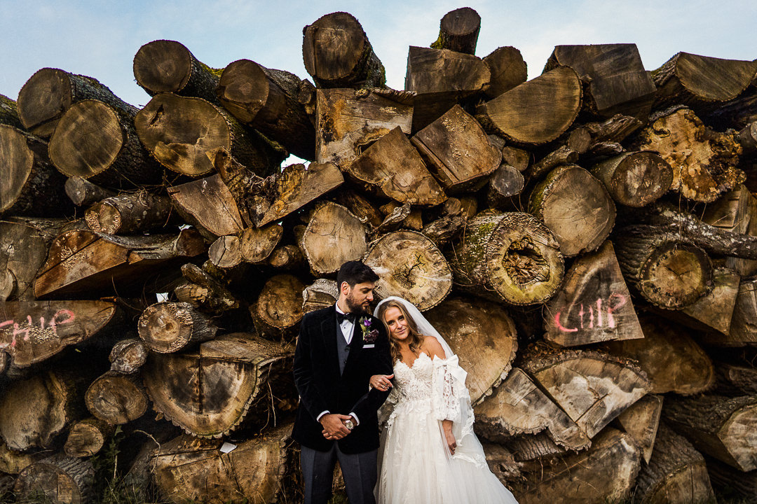A couple pose in front of a large log pile at the cotswold wedding venue Cripps barn